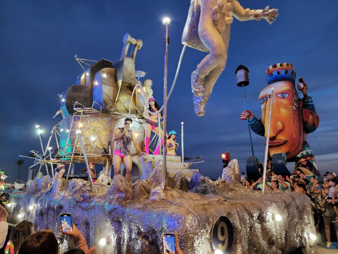 A towering, colorful illuminated parade float moving past oceanfront crowds